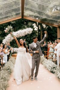 A joyful couple celebrates their wedding under a glass roof surrounded by guests.