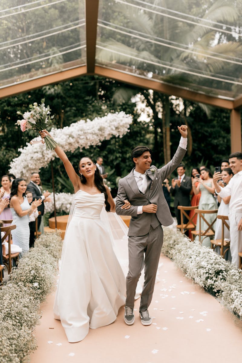 A joyful couple celebrates their wedding under a glass roof surrounded by guests.